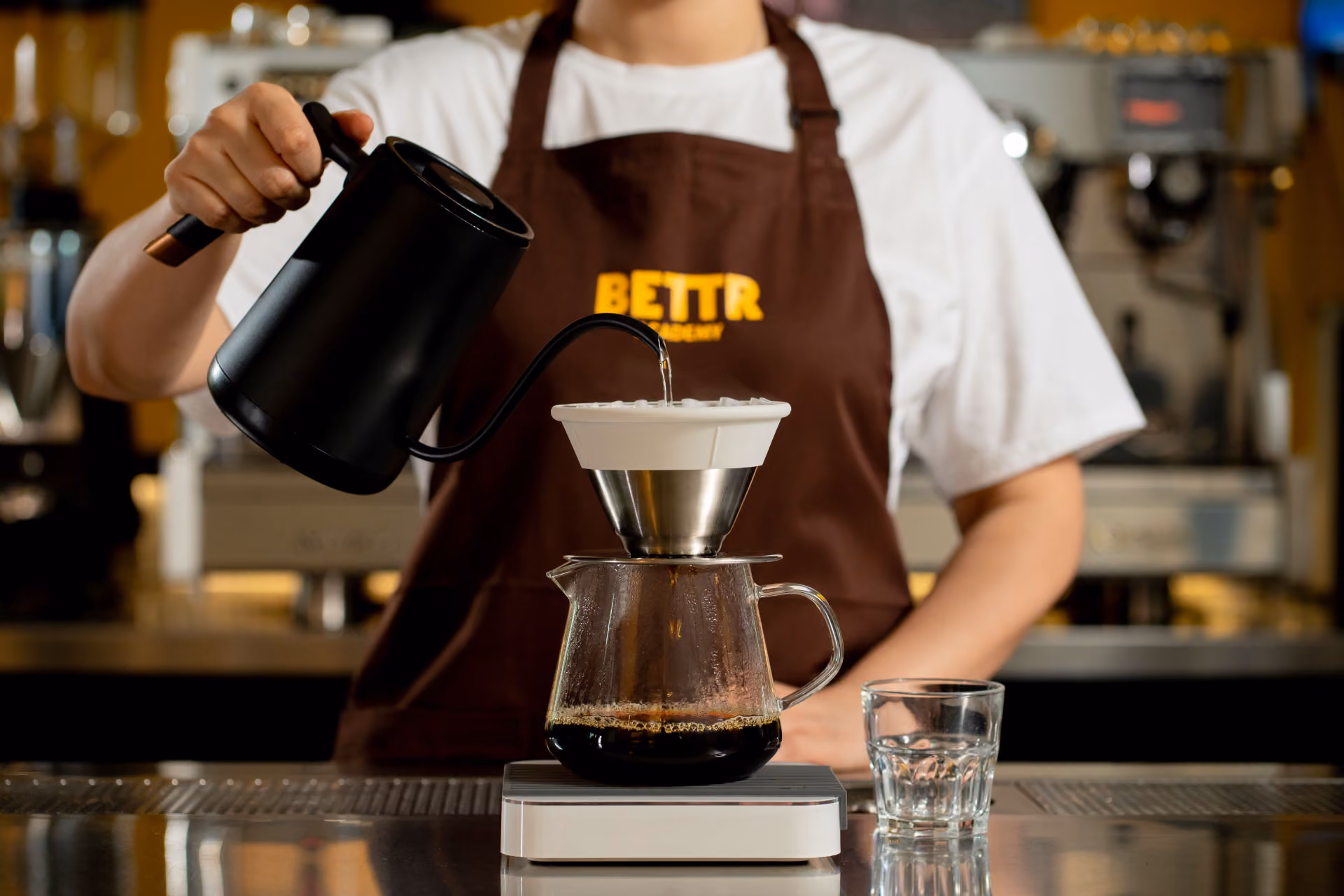 Hands holding spoons, tasting multiple cups of black coffee on a metal table.