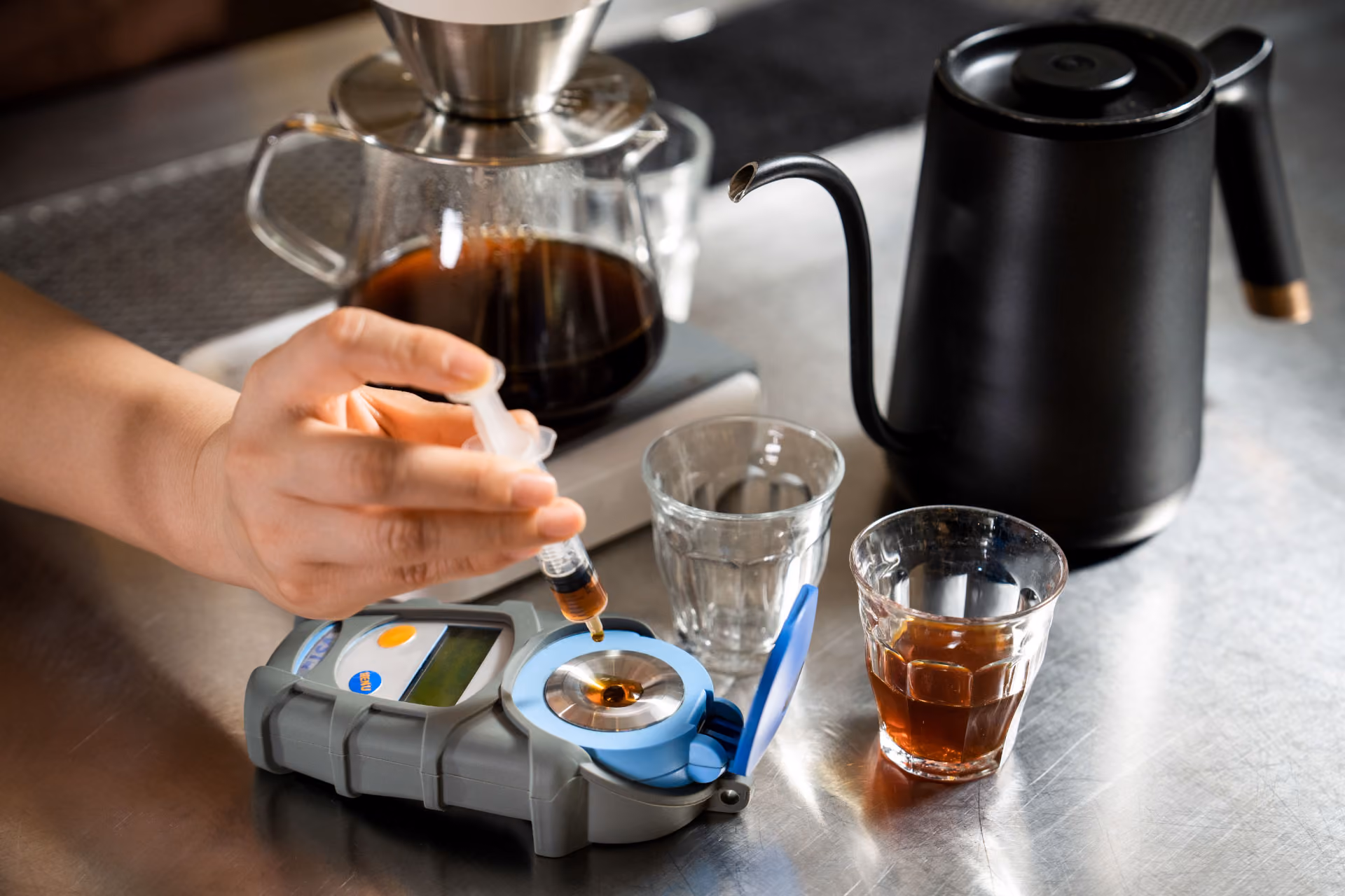 Hands holding spoons, tasting multiple cups of black coffee on a metal table.