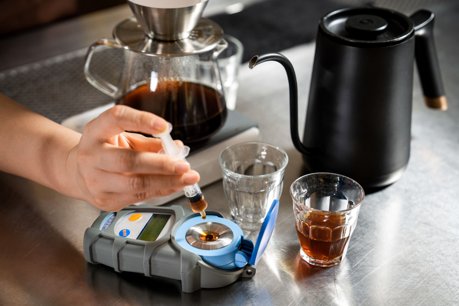 Hands holding spoons, tasting multiple cups of black coffee on a metal table.