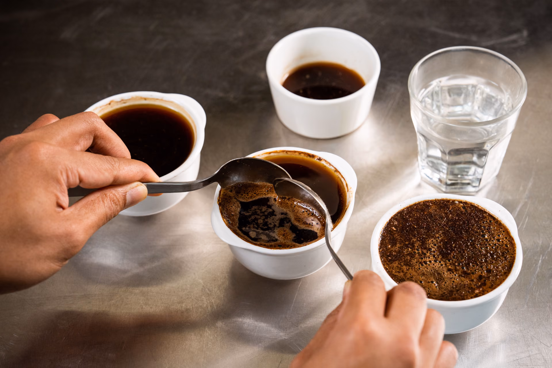 Hands holding spoons, tasting multiple cups of black coffee on a metal table.