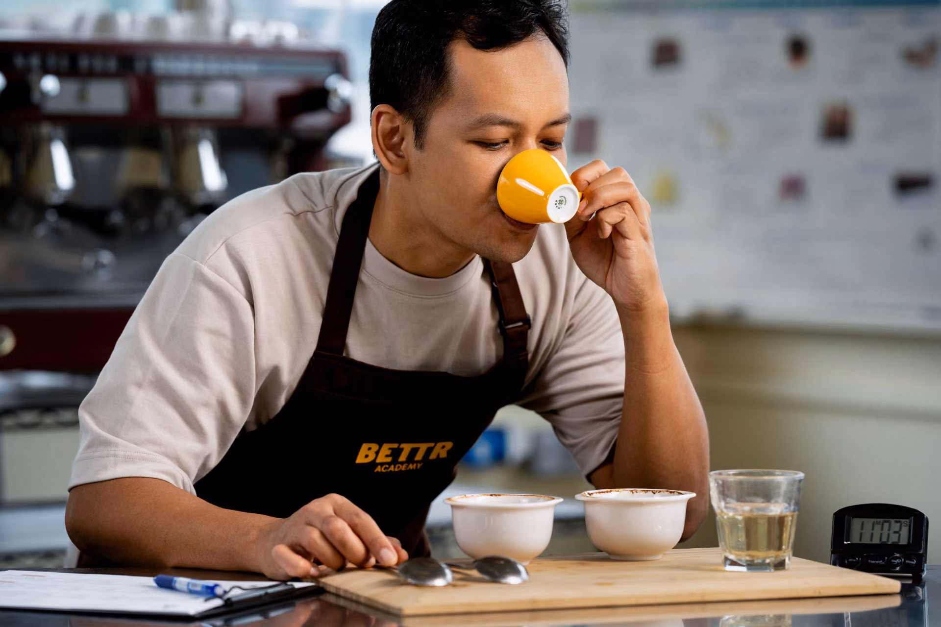 Hands holding spoons, tasting multiple cups of black coffee on a metal table.