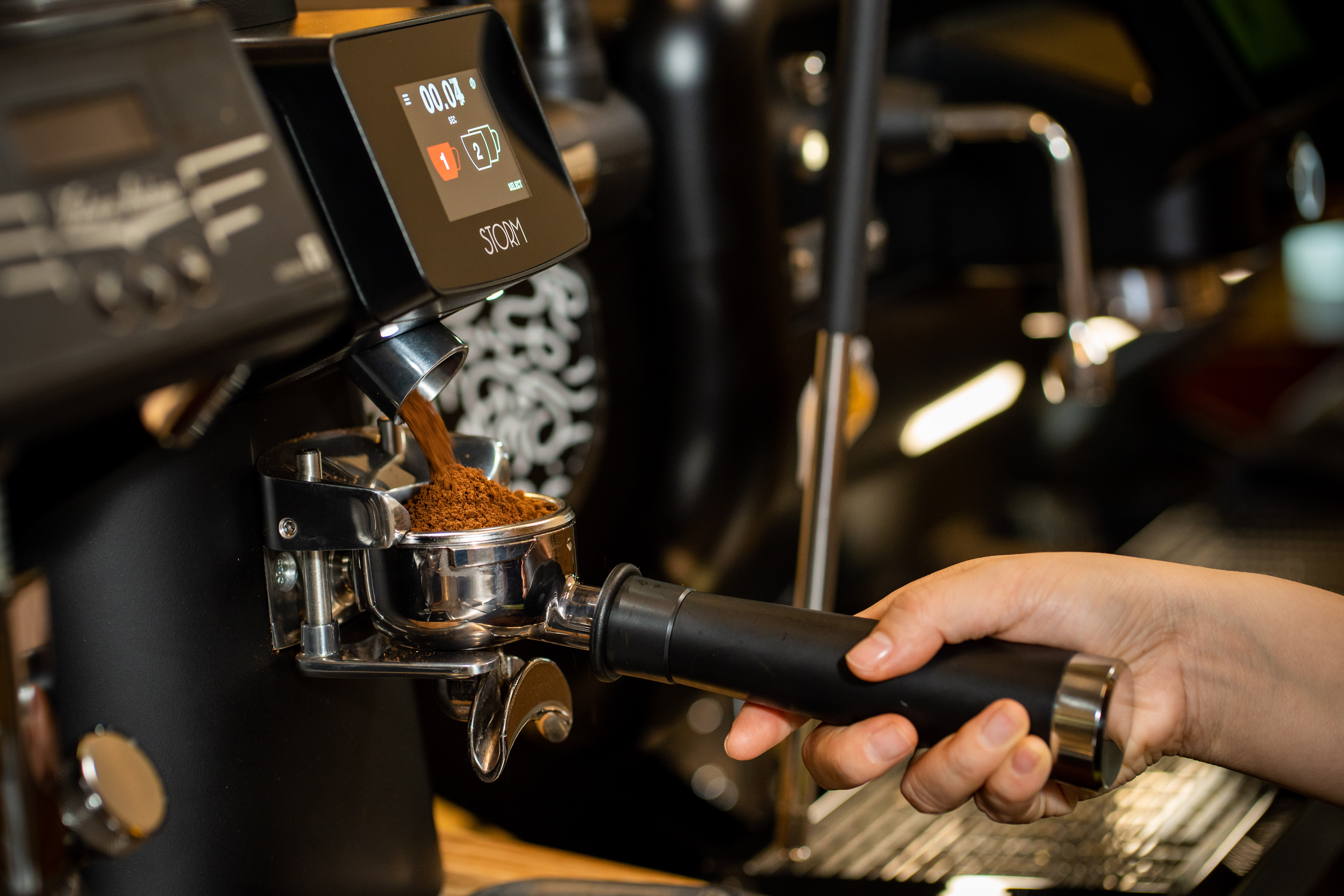 Hands holding spoons, tasting multiple cups of black coffee on a metal table.