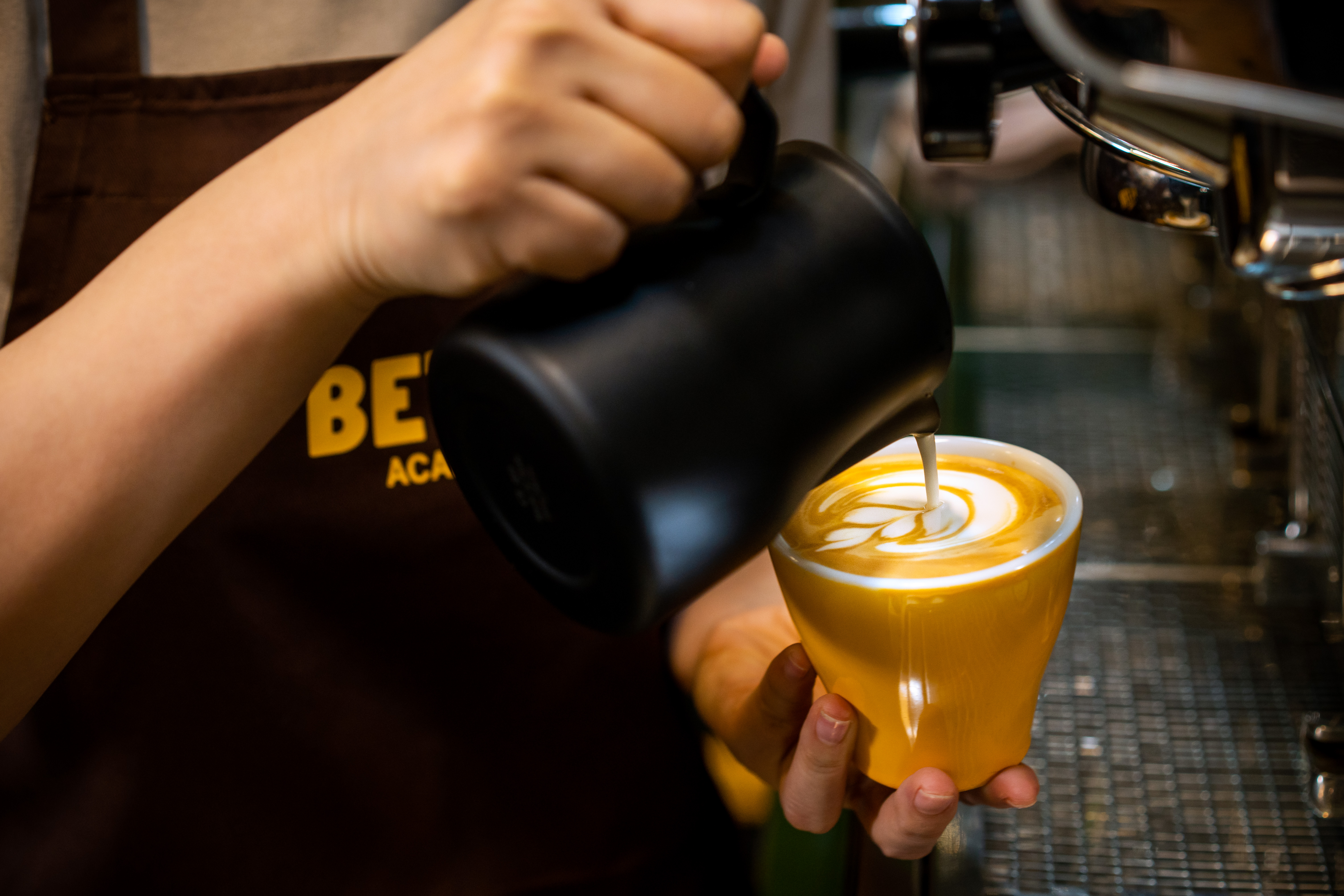 Hands holding spoons, tasting multiple cups of black coffee on a metal table.