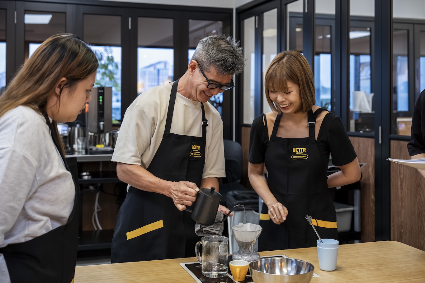 Hands holding spoons, tasting multiple cups of black coffee on a metal table.