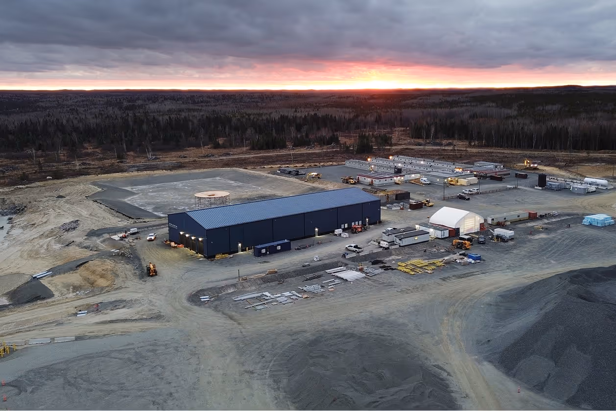 Aerial view of industrial site with buildings, vehicles, and equipment at sunset near a forest.