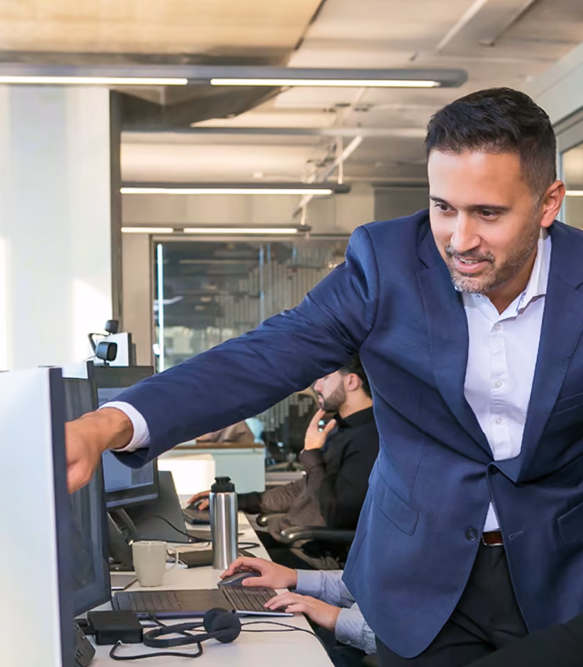 Man in blue blazer pointing at computer screen while another person types on a keyboard in office.