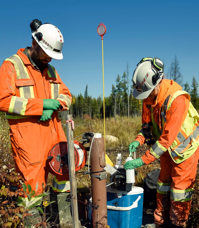 Two workers in orange safety suits and helmets handling equipment outdoors near grass and trees.