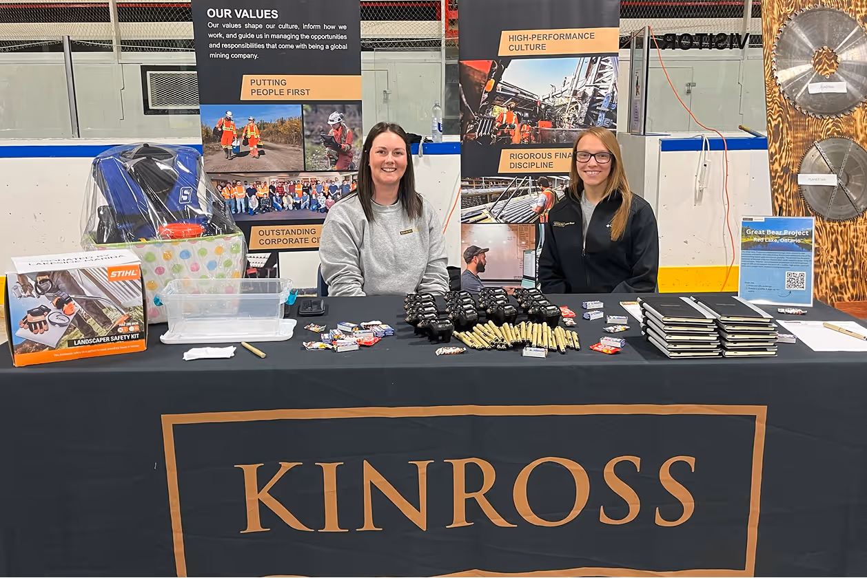 Two women sit behind a Kinross-branded table with promotional items and informational banners.