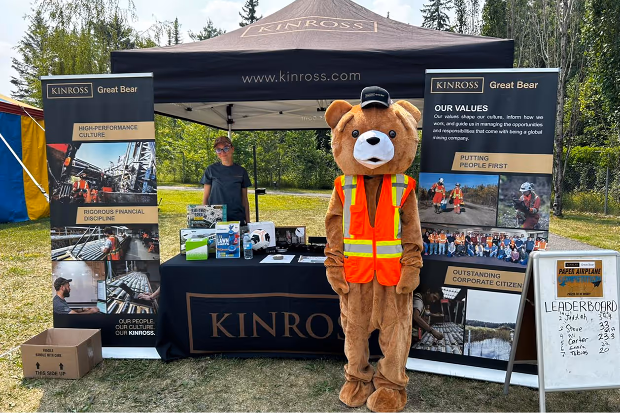 Person in bear costume wearing orange safety vest stands in front of Kinross Great Bear booth at outdoor event.