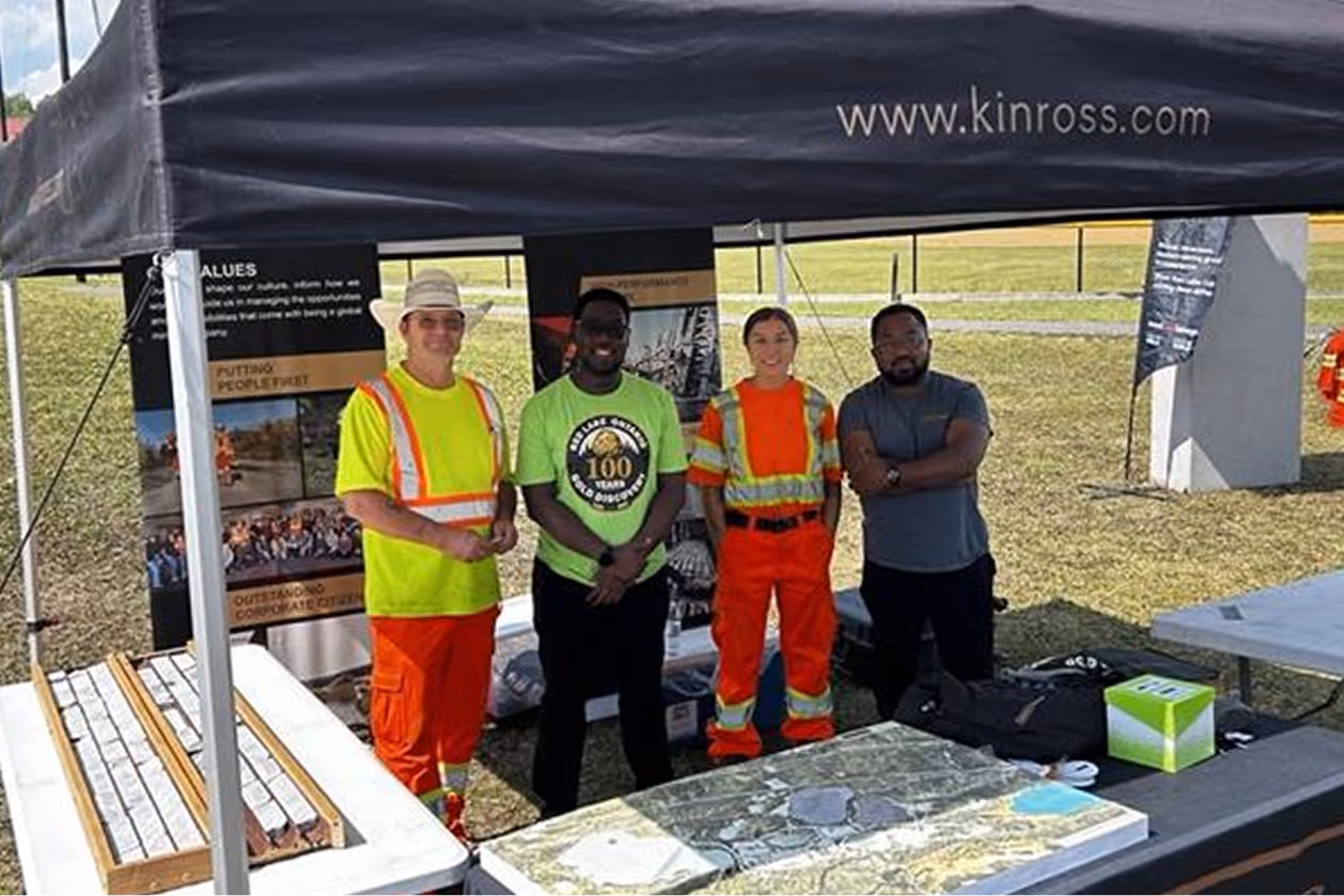 Four people stand under a tent with kinross.com above, next to tables with maps and samples outdoors.