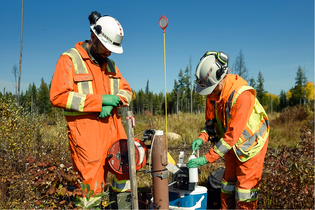 Two workers in orange safety gear and helmets collecting samples in a forested field area.