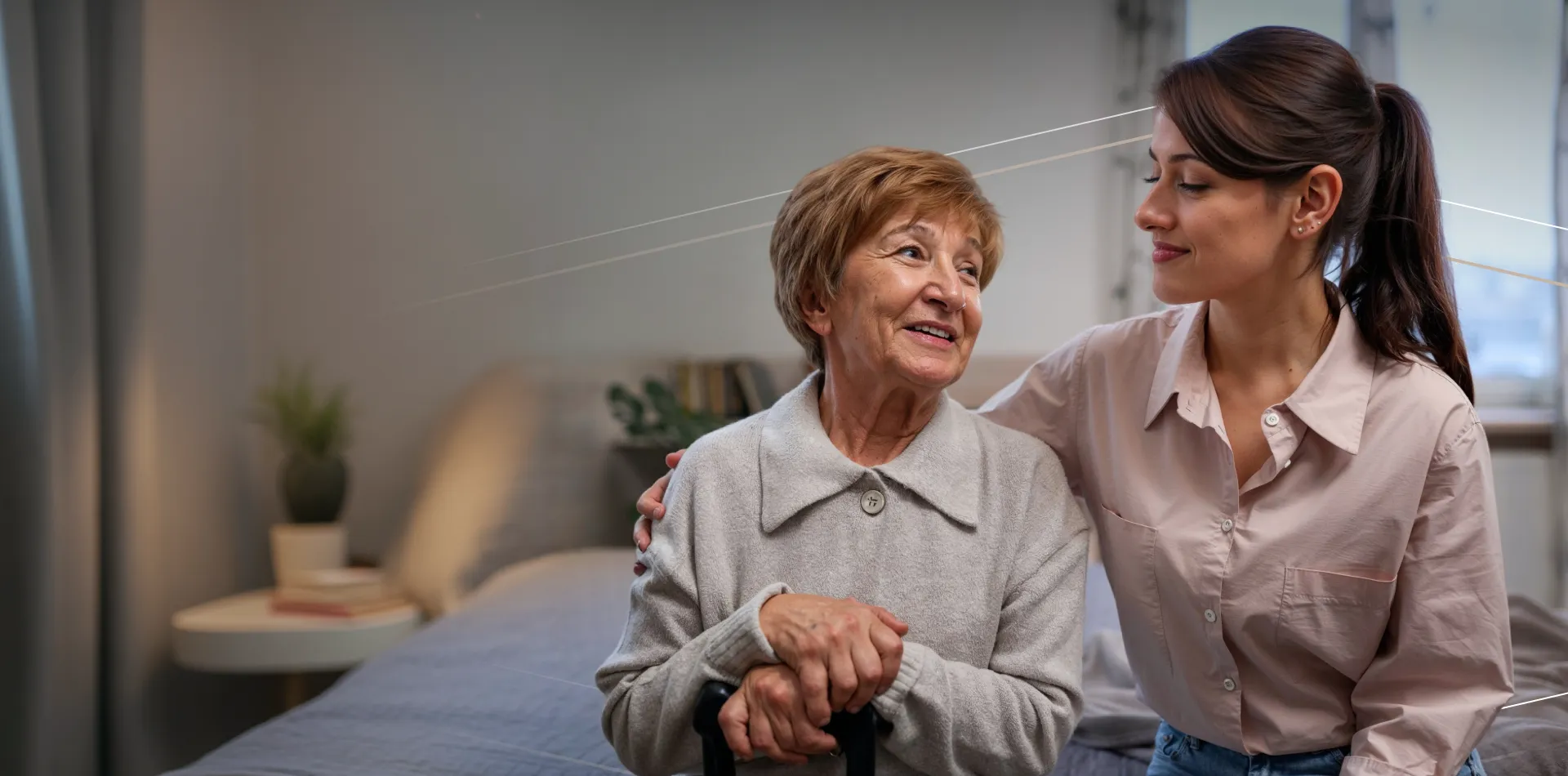 Young woman gently supporting and smiling at an elderly woman sitting on a bed in a cozy room.