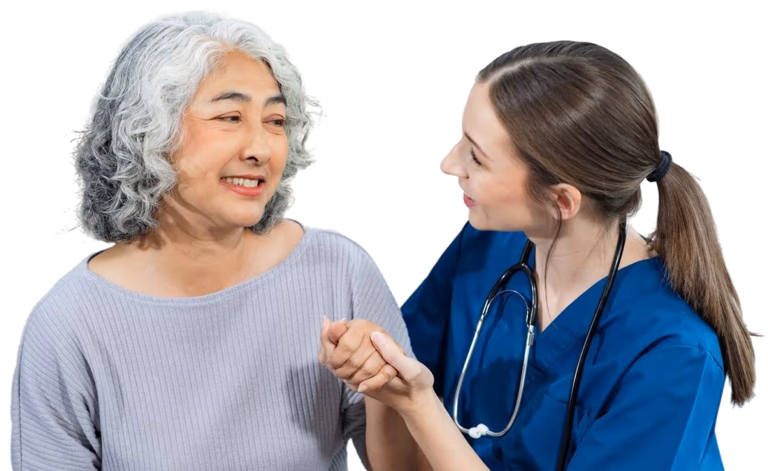 Nurse in scrubs holding hands and smiling at an older woman with gray curly hair in a gray shirt.