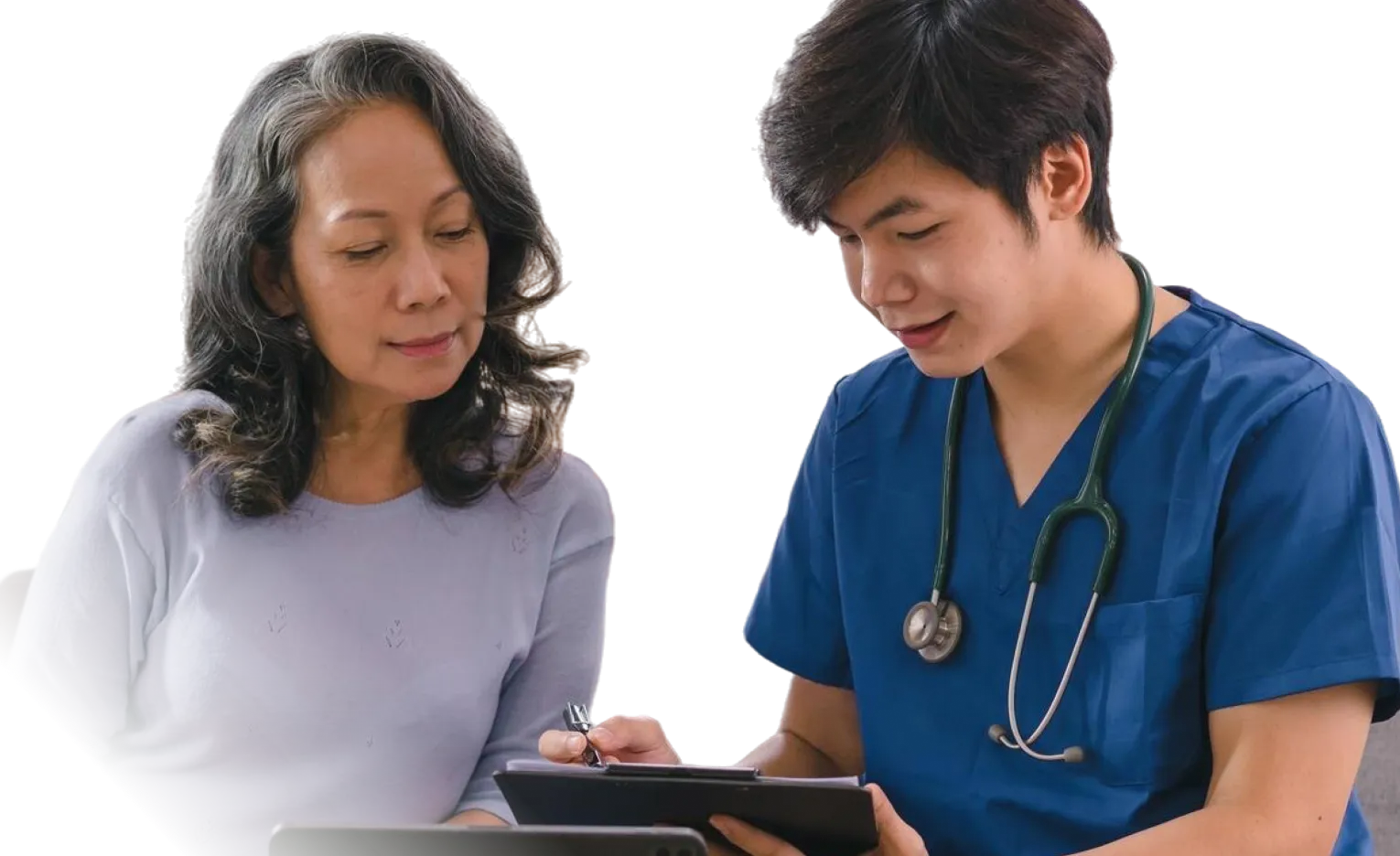 Male nurse in blue scrubs with stethoscope discussing notes with middle-aged woman in lavender sweater.