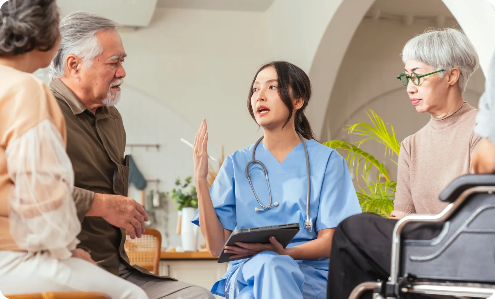 Female nurse in blue scrubs talking to elderly people, one in a wheelchair, during a consultation.