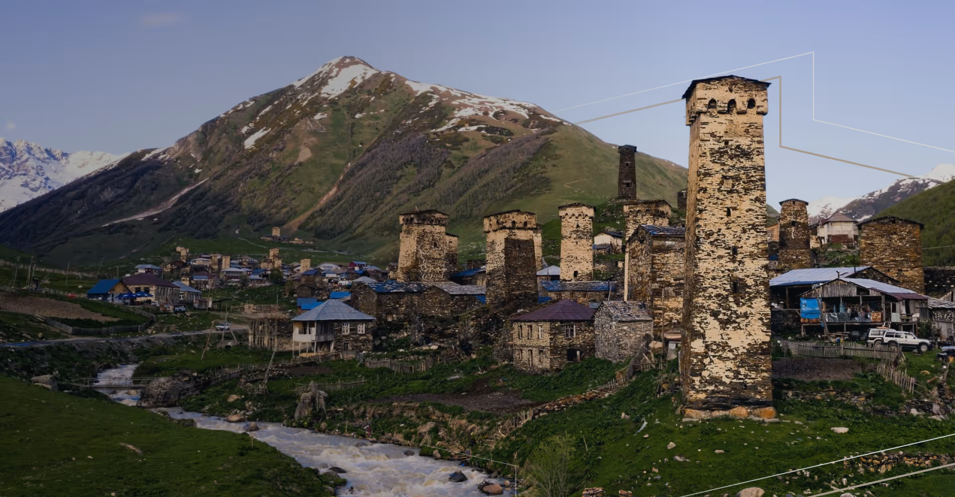 Ancient stone towers and houses in a green valley with a flowing river and snow-capped mountains behind.