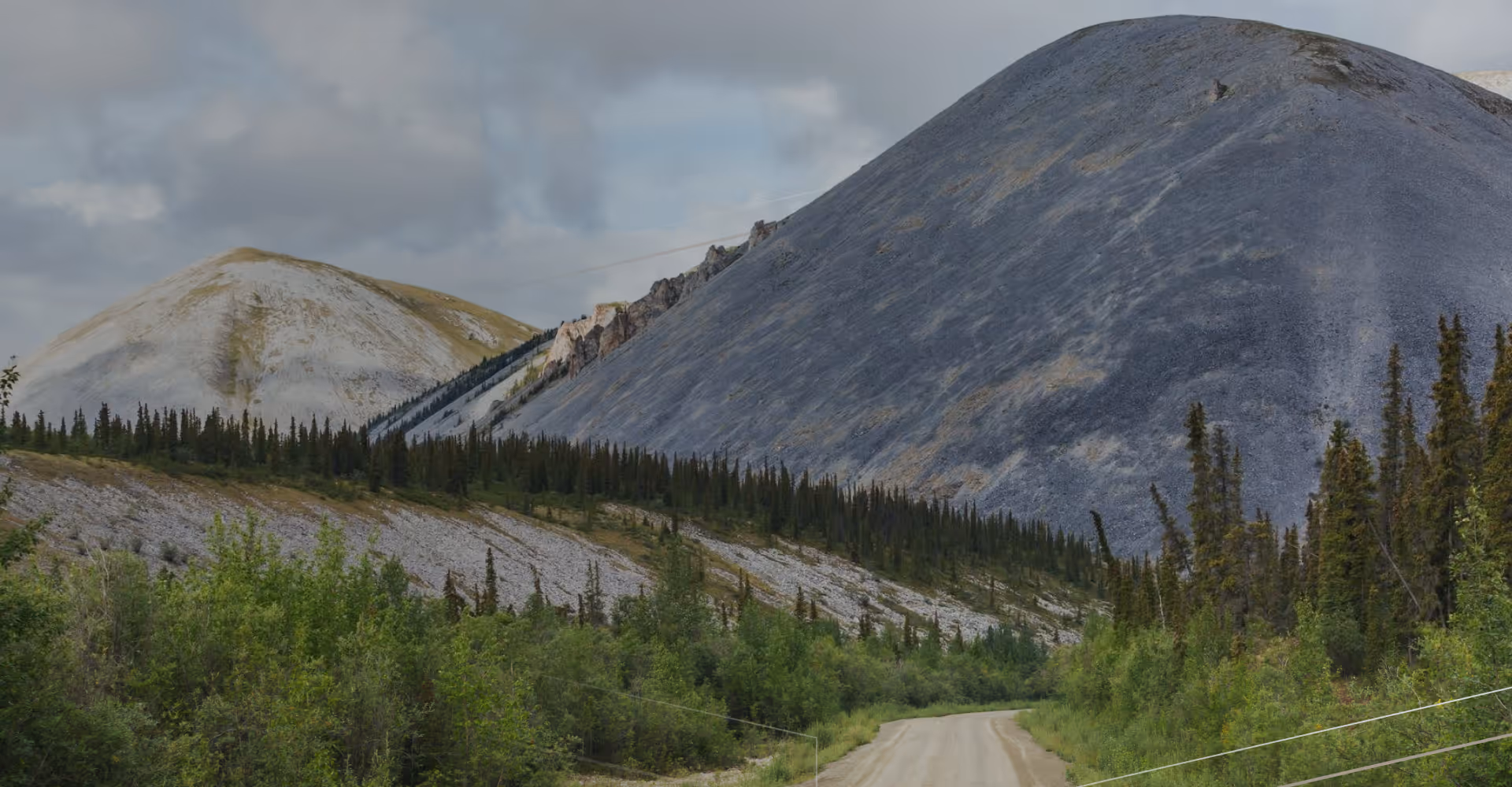 Dirt road winds through green forest with steep rocky mountains and cloudy sky in the background.