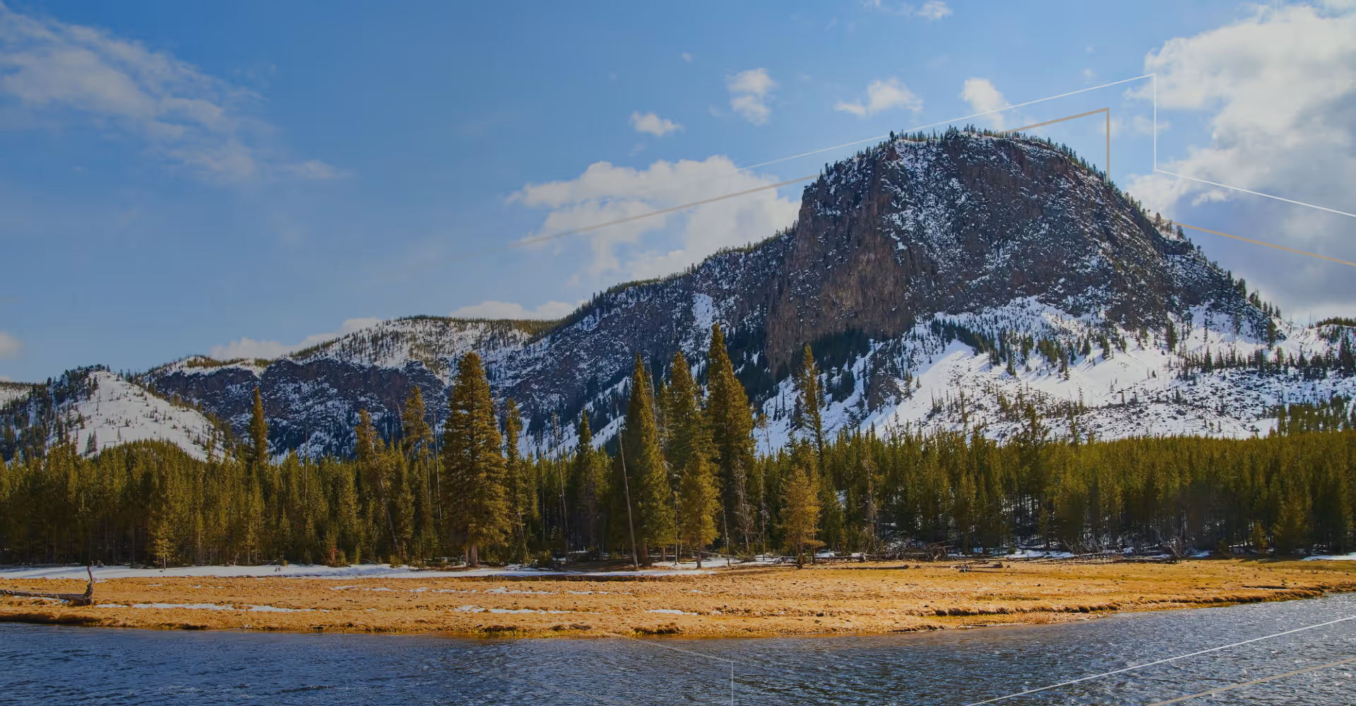 Snow-capped mountain overlooking pine forest and grassy shoreline by a calm river under blue sky.