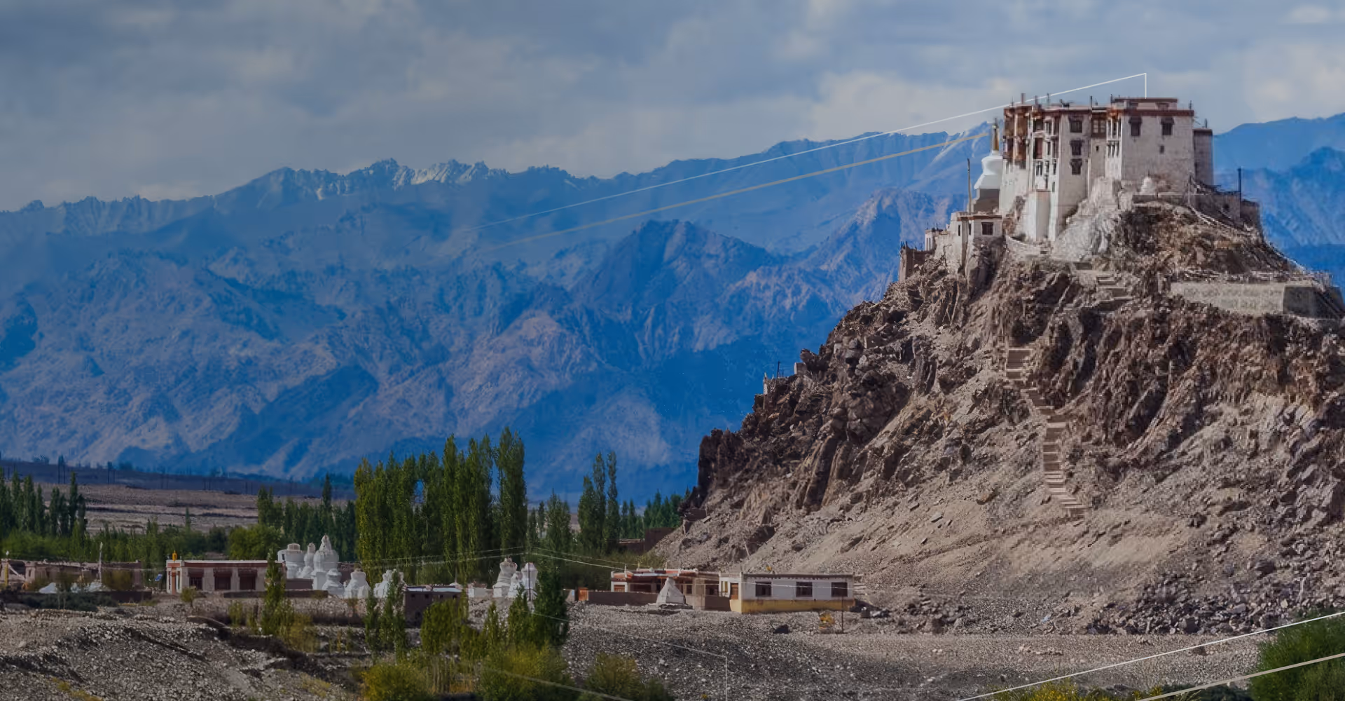 Ancient monastery perched on rocky hill with green trees and mountain range in the background.