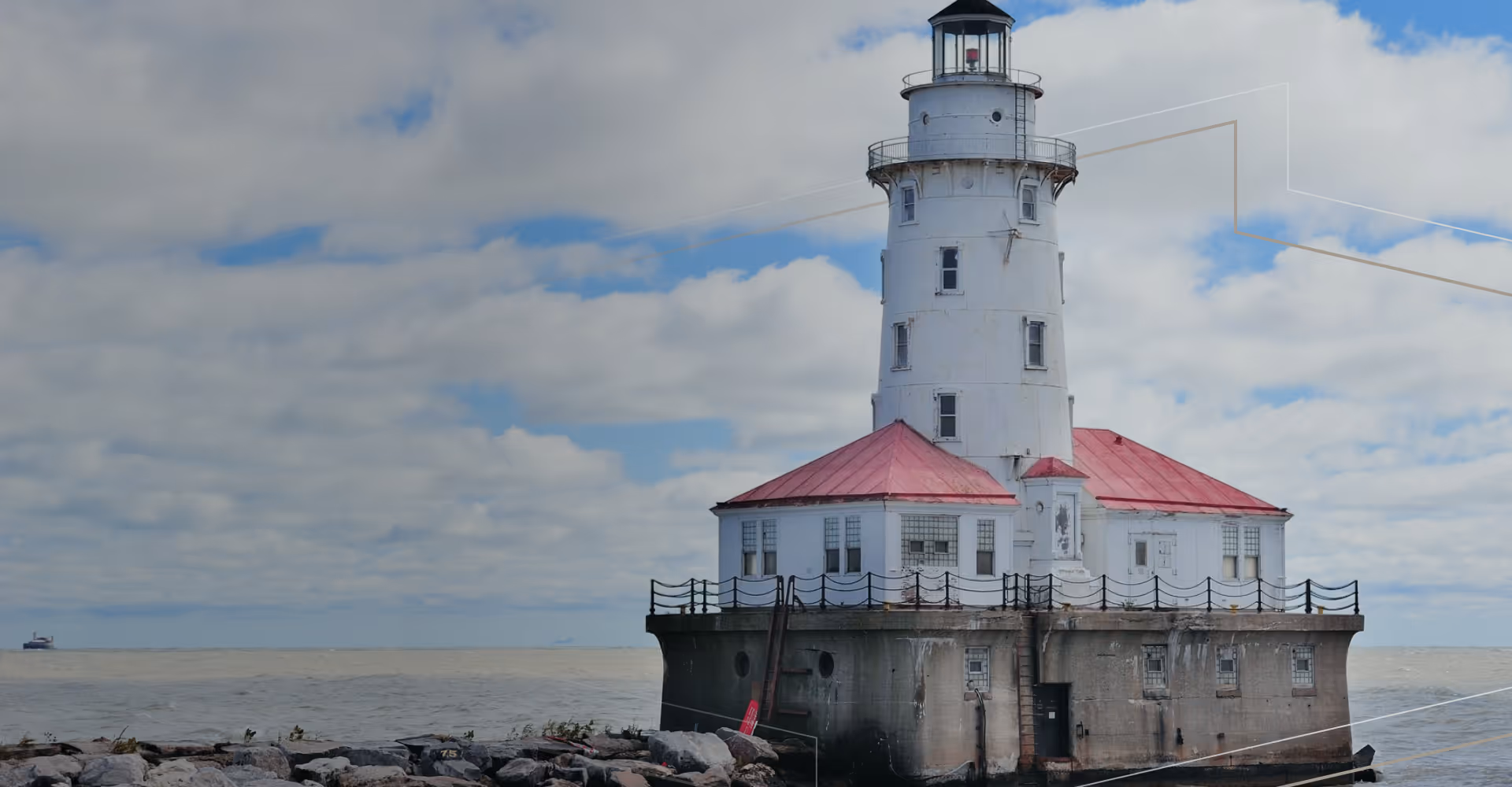 White lighthouse with red roof on a concrete base by a rocky shoreline under a cloudy sky.