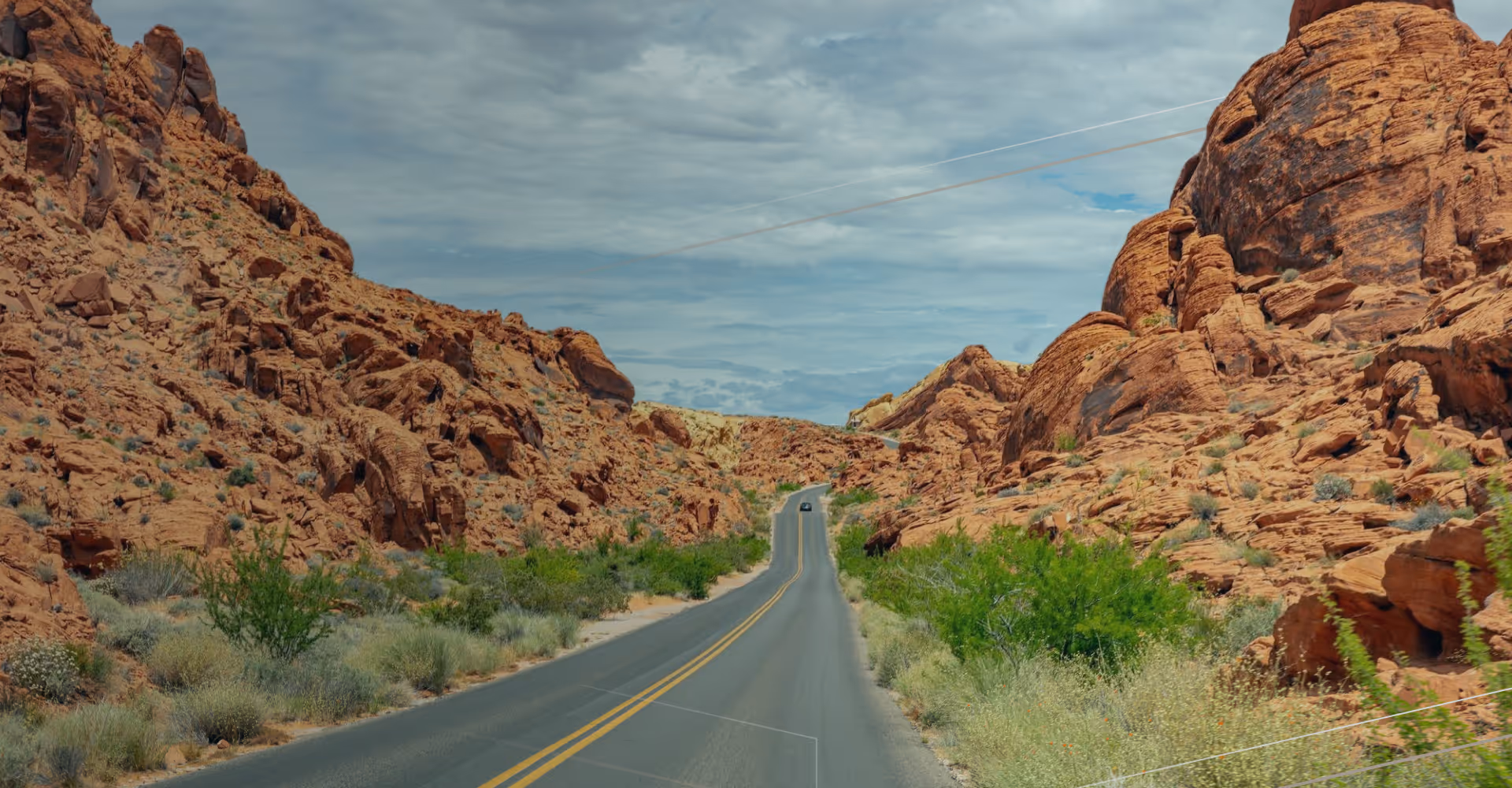 Two-lane road through rocky red desert hills with green shrubs under a cloudy sky.