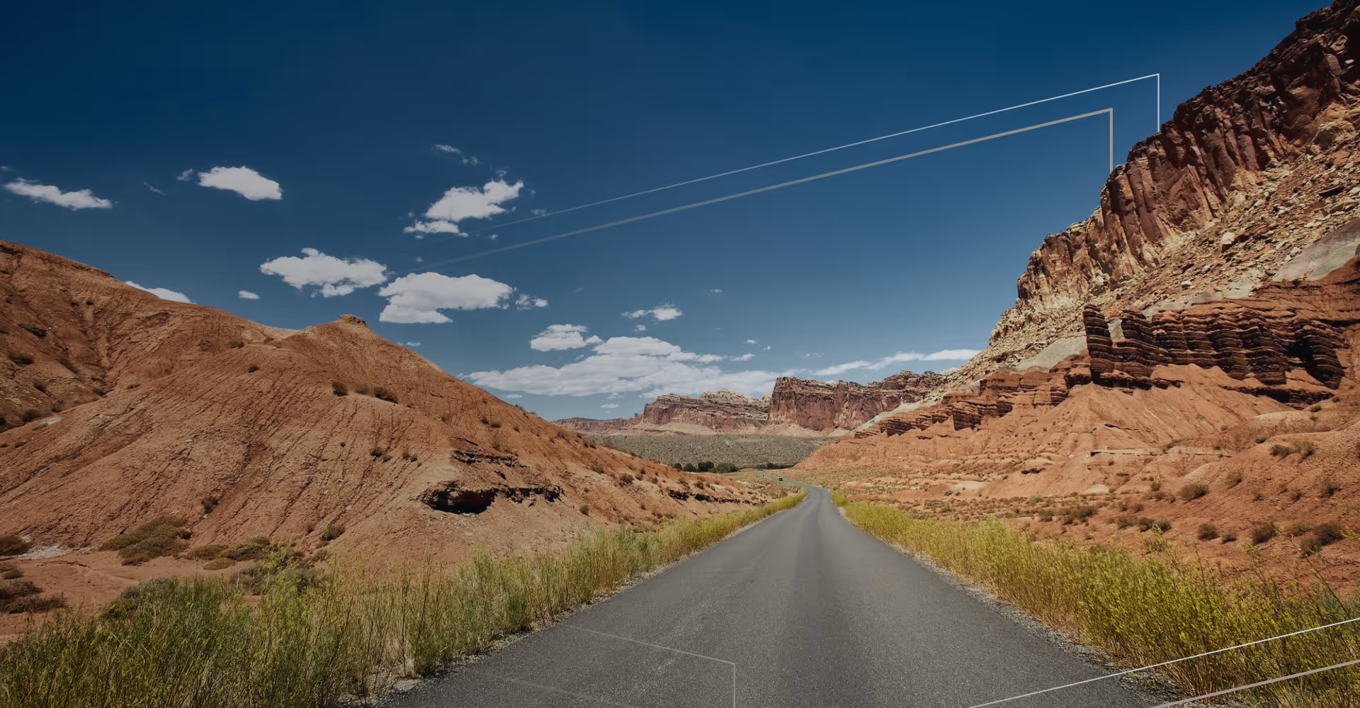 Asphalt road leading through red rock formations under a blue sky with scattered clouds.
