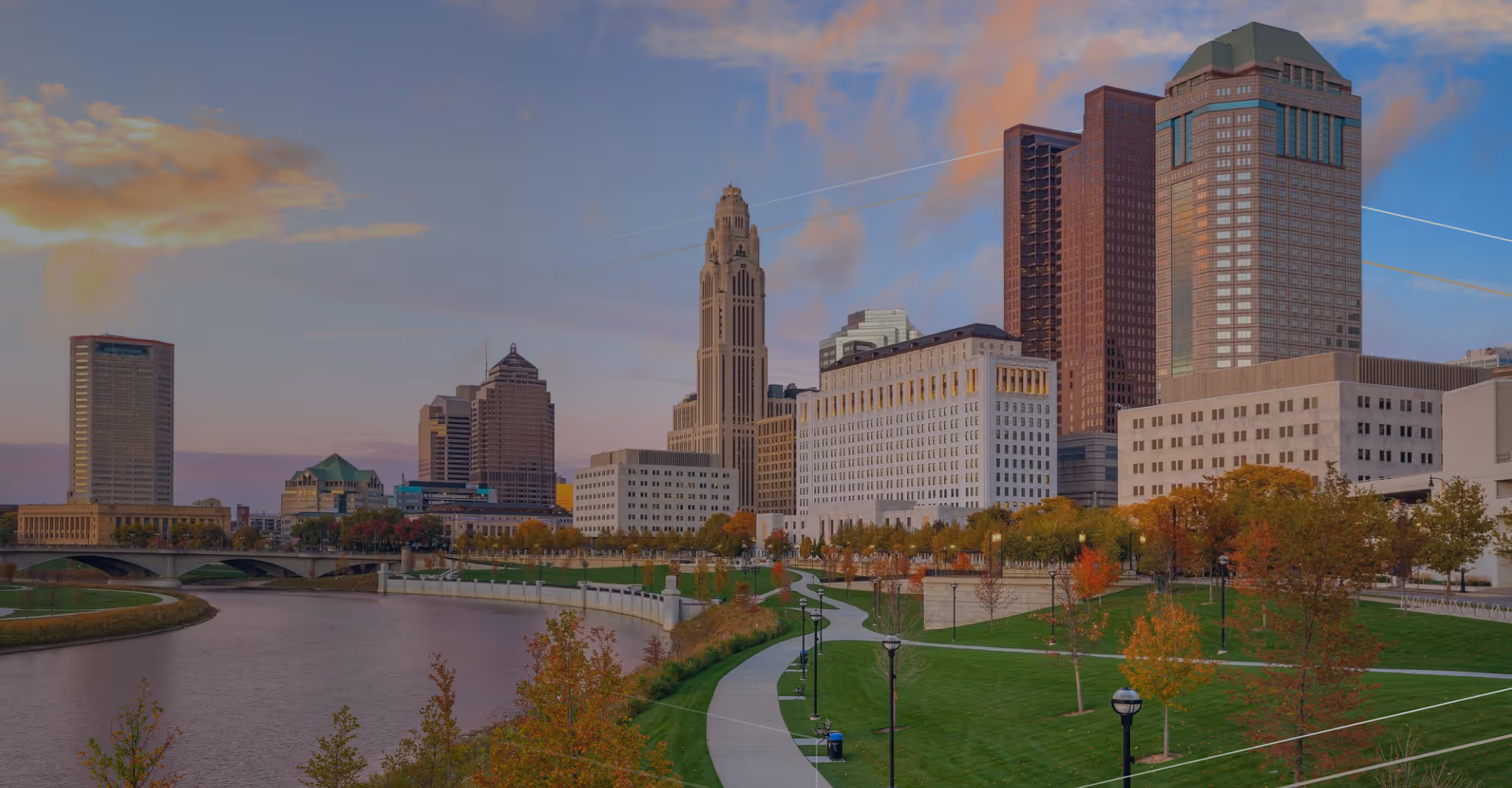 Riverfront park with fall trees and city skyline under colorful evening sky.