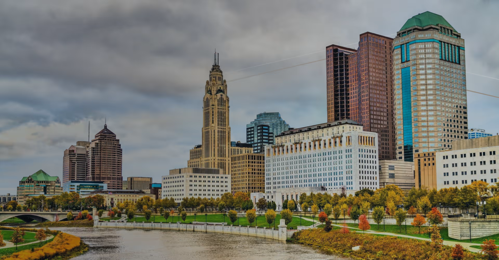City skyline with diverse buildings and autumn trees along a river under a cloudy sky.