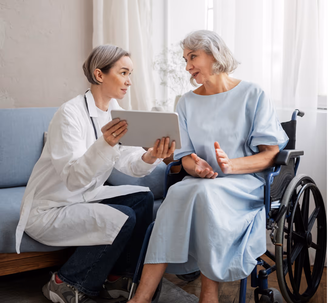 Doctor showing tablet to an elderly woman in a wheelchair during a consultation indoors.