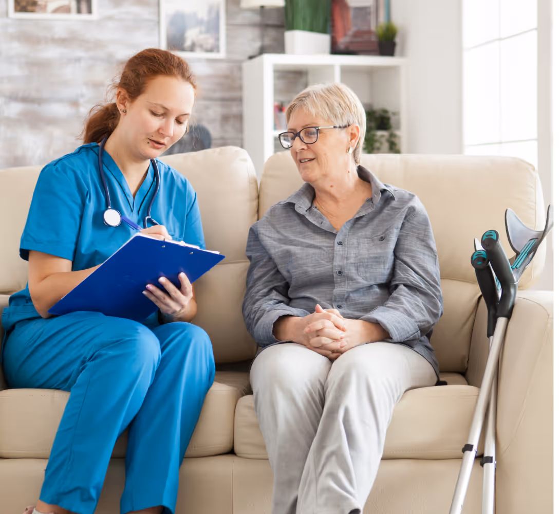 Healthcare worker in blue scrubs with clipboard talks to seated elderly woman with crutches nearby.