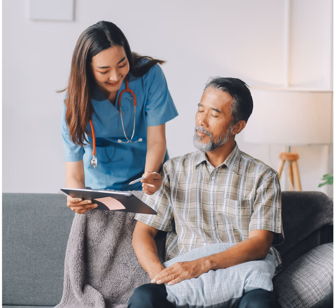 Nurse with stethoscope showing a clipboard to an elderly man seated on a couch.