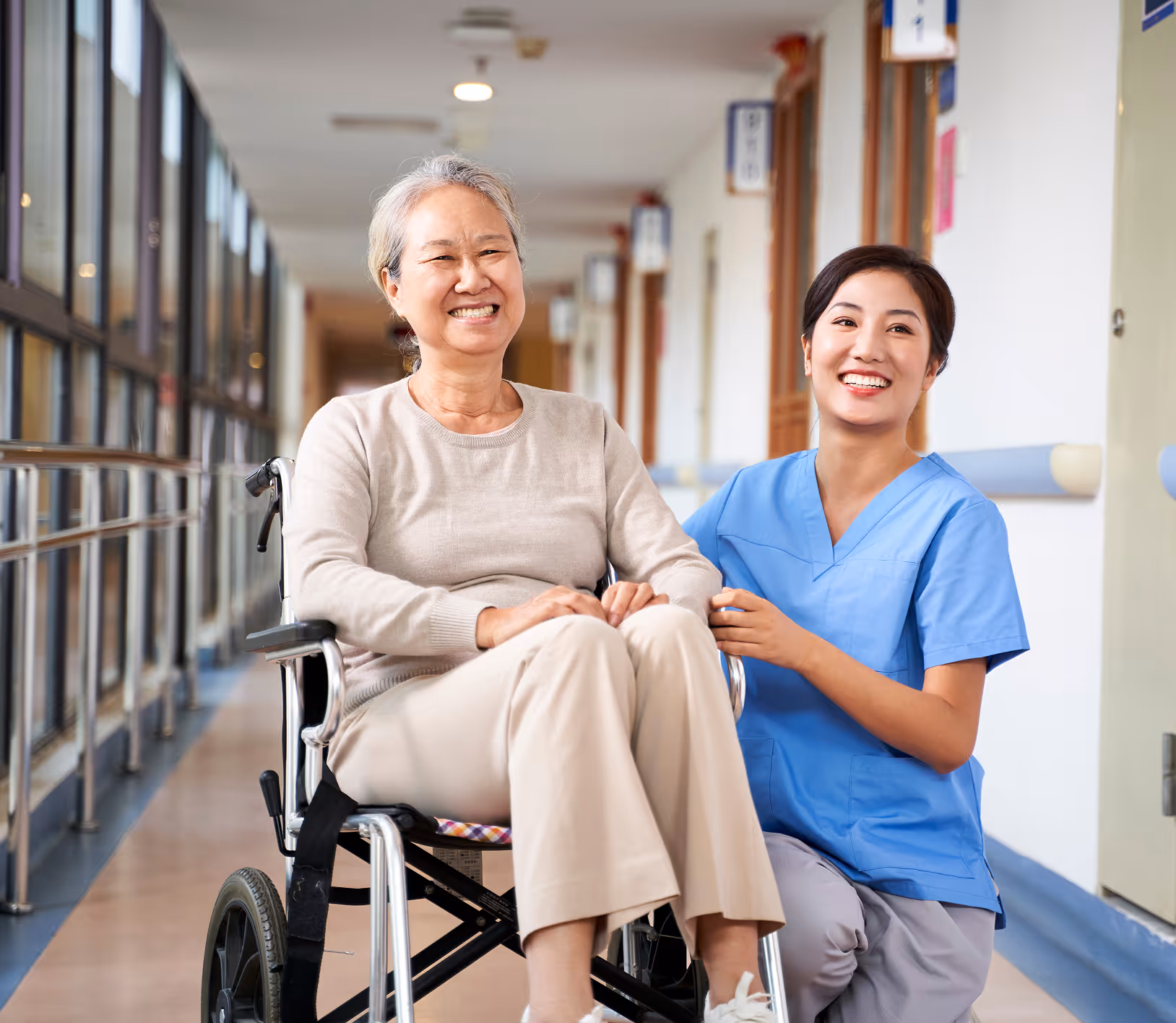 Smiling nurse in blue scrubs kneeling beside a happy elderly woman sitting in a wheelchair in a hallway.