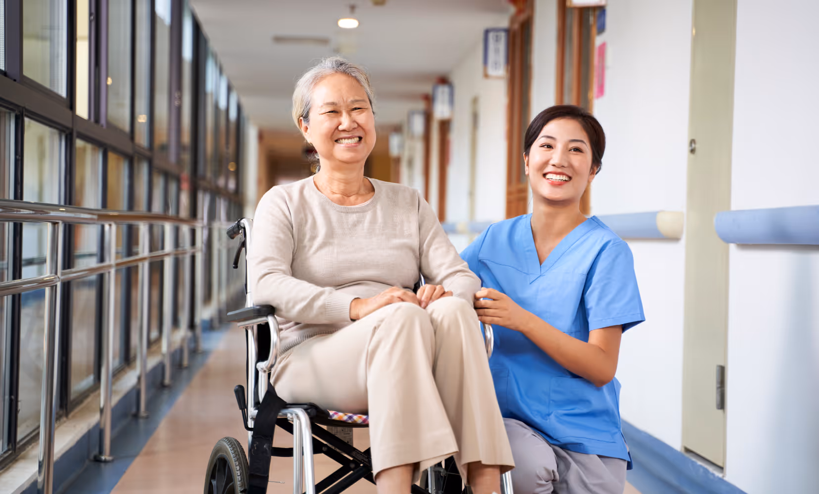 Smiling nurse kneels beside happy elderly woman sitting in wheelchair in a bright hallway.