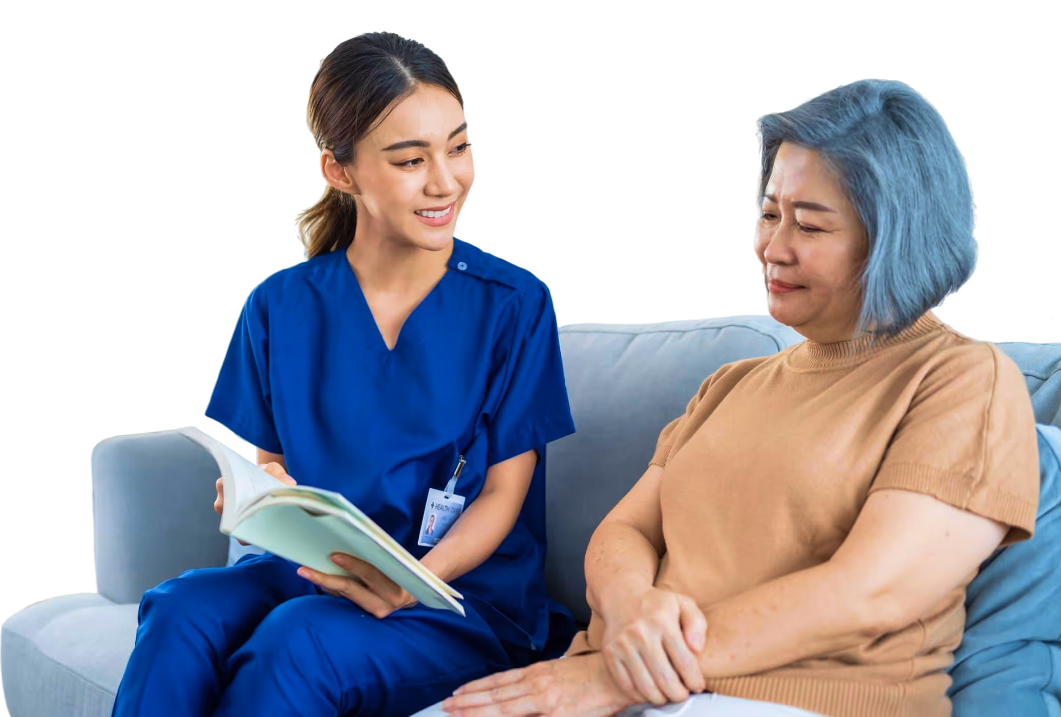 Healthcare worker in blue scrubs talking to an elderly woman sitting on a couch.