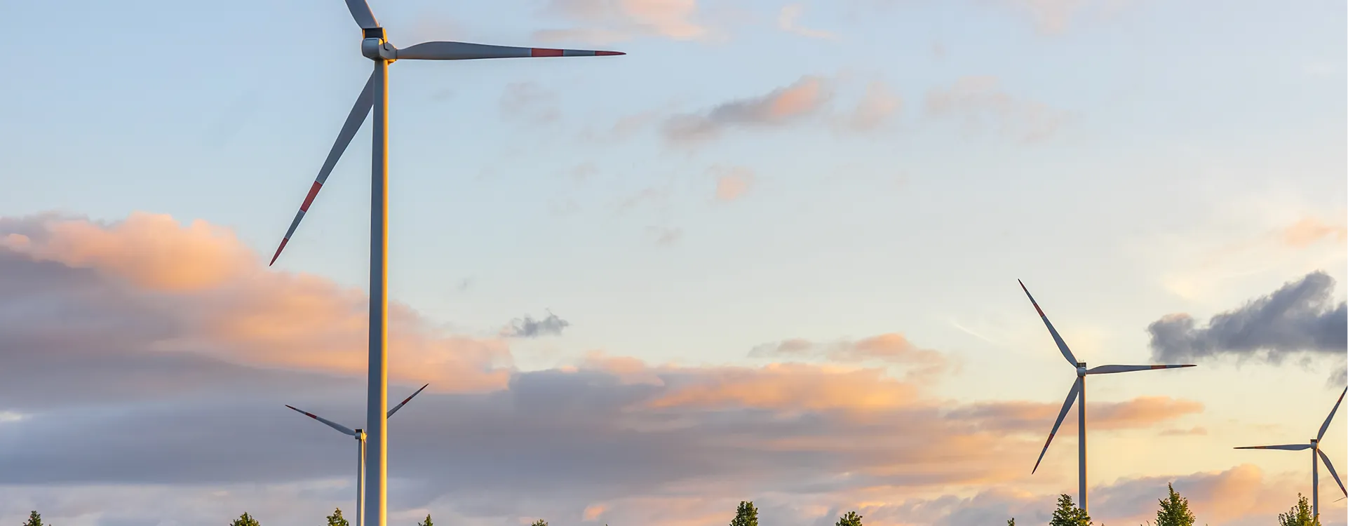 Wind turbines in a field under a colorful sunset sky with scattered clouds and trees below.