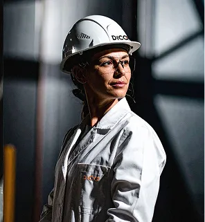 Woman engineer in white coat and hard hat looking to side against industrial background.
