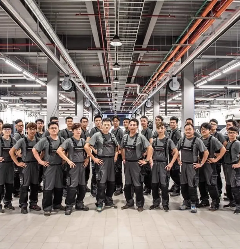 Group of factory workers wearing gray uniforms standing in a large industrial facility.