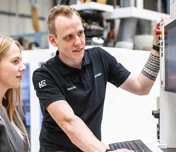 Man with tattoo teaching woman how to use industrial machine control panel.