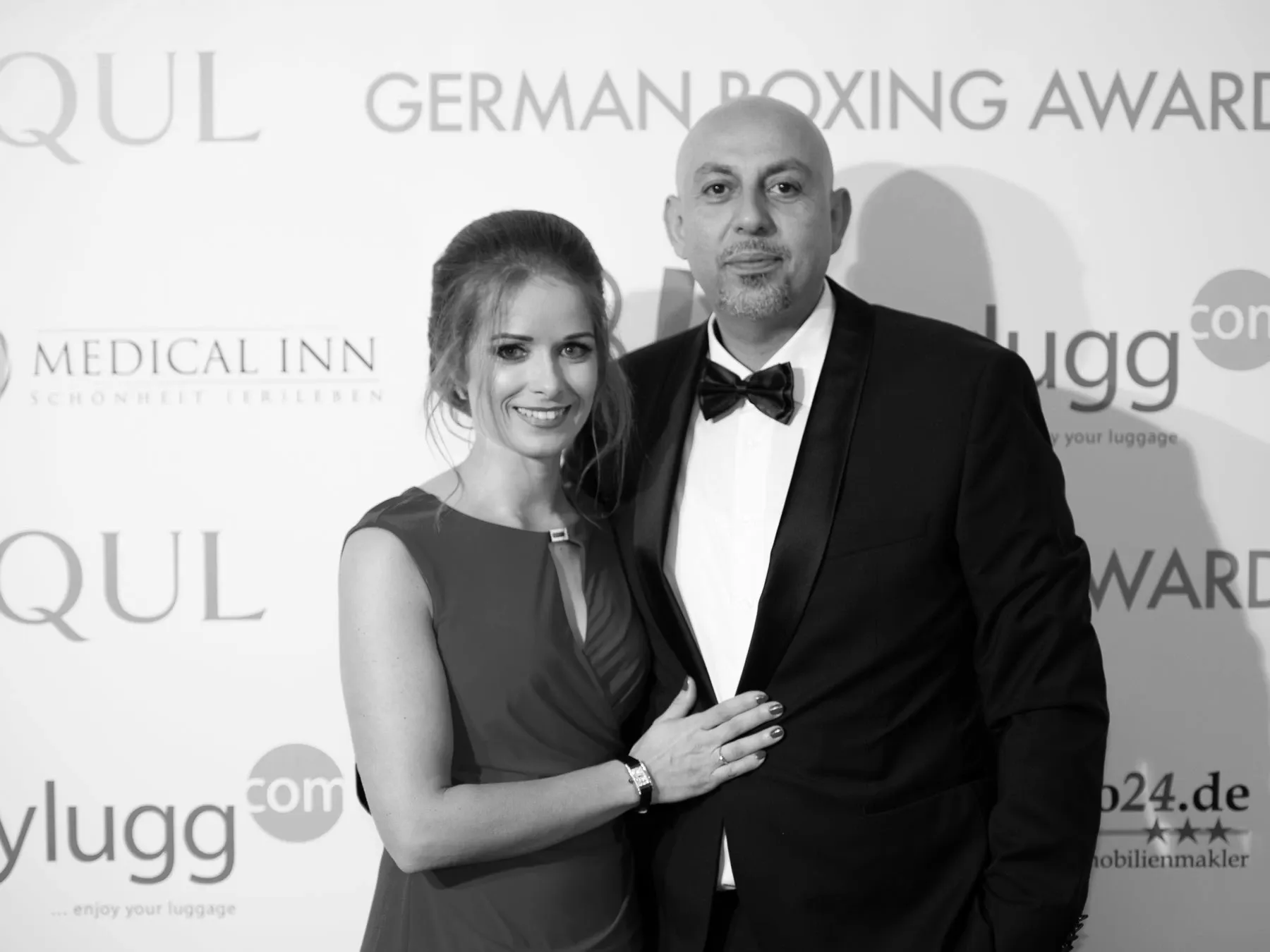 Woman in sleeveless dress smiling with hand on chest of man in tuxedo and bow tie at event backdrop.