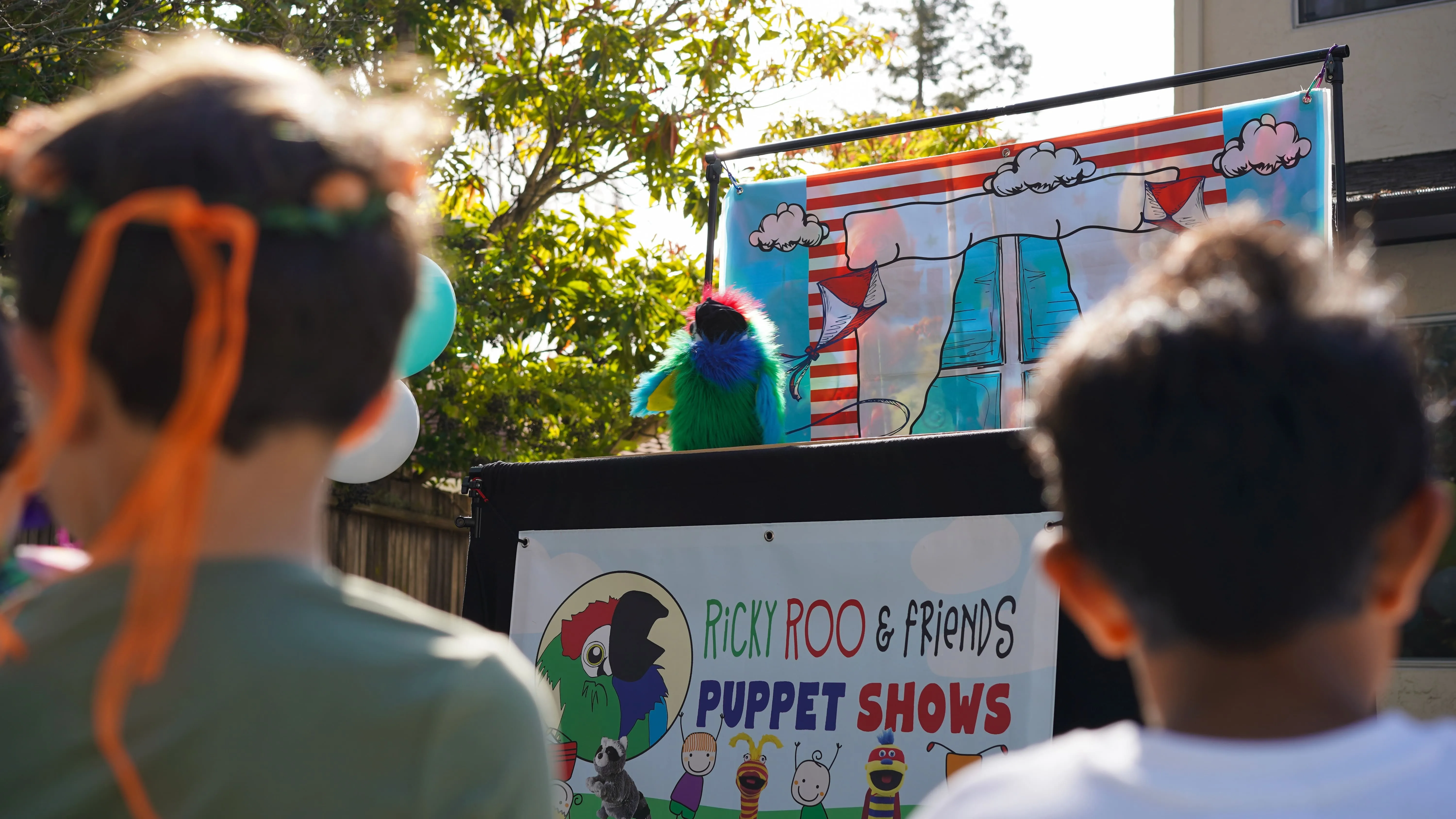 Two children watch a colorful parrot puppet in a Ricky Roo & Friends puppet show outdoors.