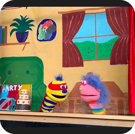Two Ricky Roo & Friends puppets with colorful stripes and hair performing in a cozy room with window, curtains, and decorations.