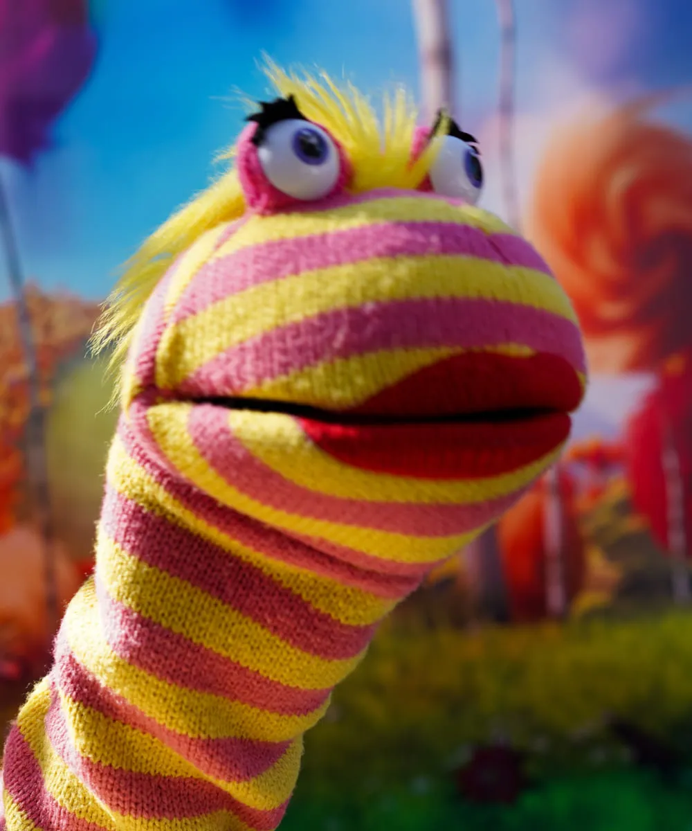 Colorful Ricky Roo puppet with large eyes and striped yellow and pink knitted fabric in a bright outdoor setting.