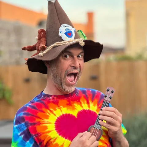 Ricky Roo & Friends founder Chris Rachowicz wearing a brown wizard hat with toys and a colorful tie-dye shirt, playing a small ukulele.