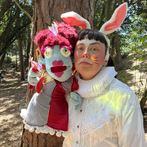 Person with bunny ears and whiskers face paint holding a colorful puppet with red curly hair.