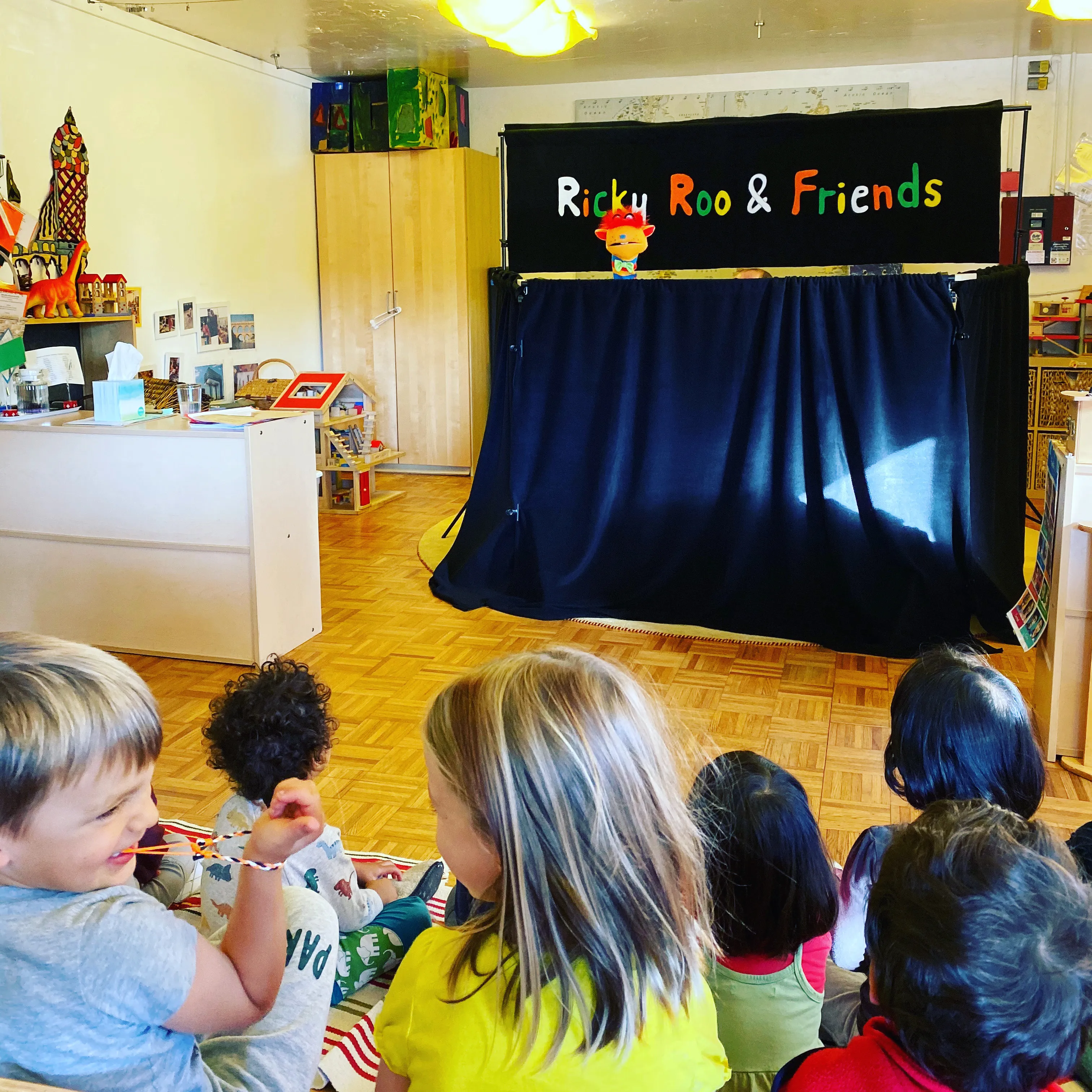 Children watching a puppet show with a colorful puppet at a theater labeled Ricky Roo & Friends.