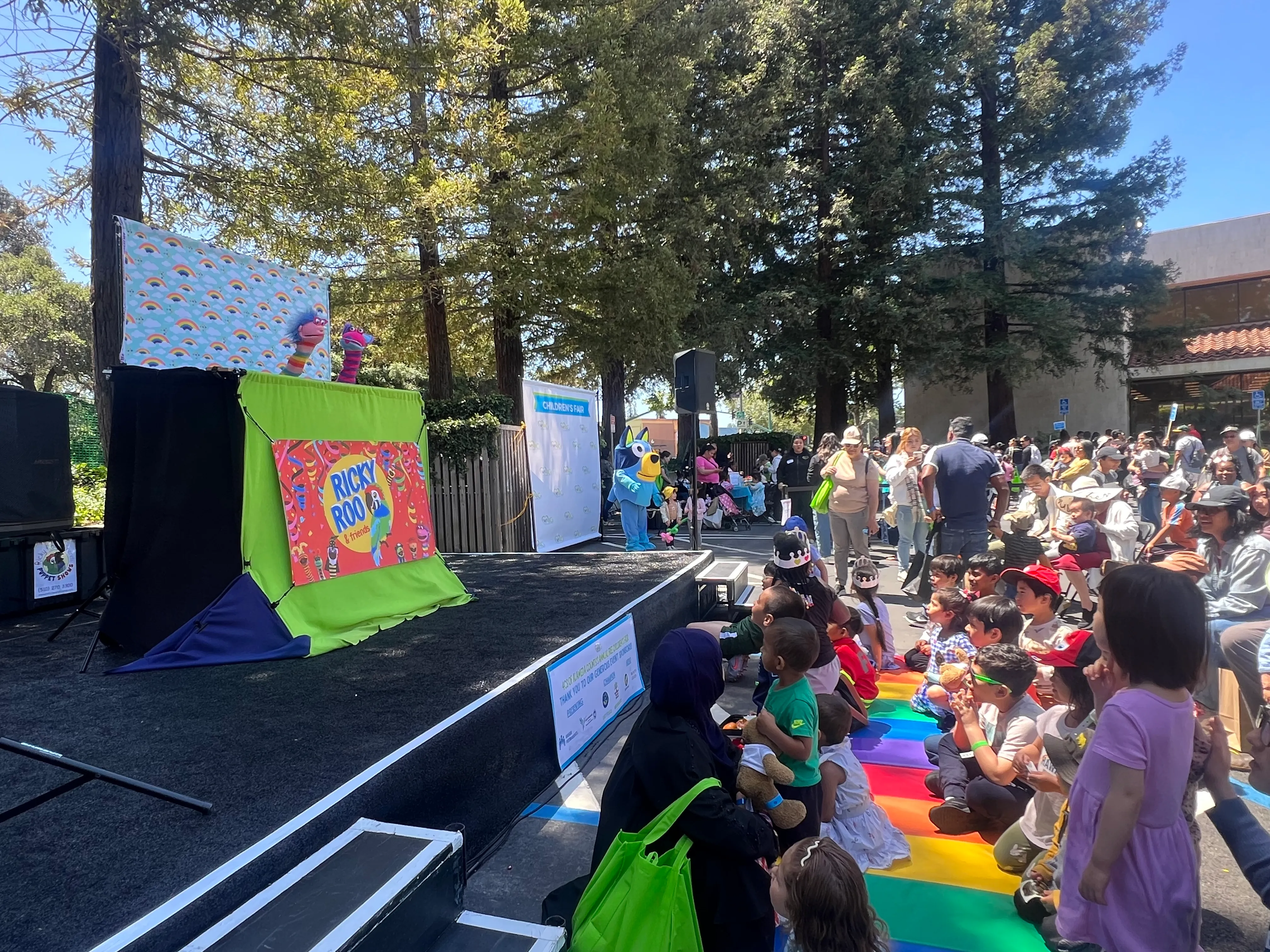 Children seated on a rainbow mat watch a Ricky Roo & Friends puppet show on an outdoor stage at a community fair.