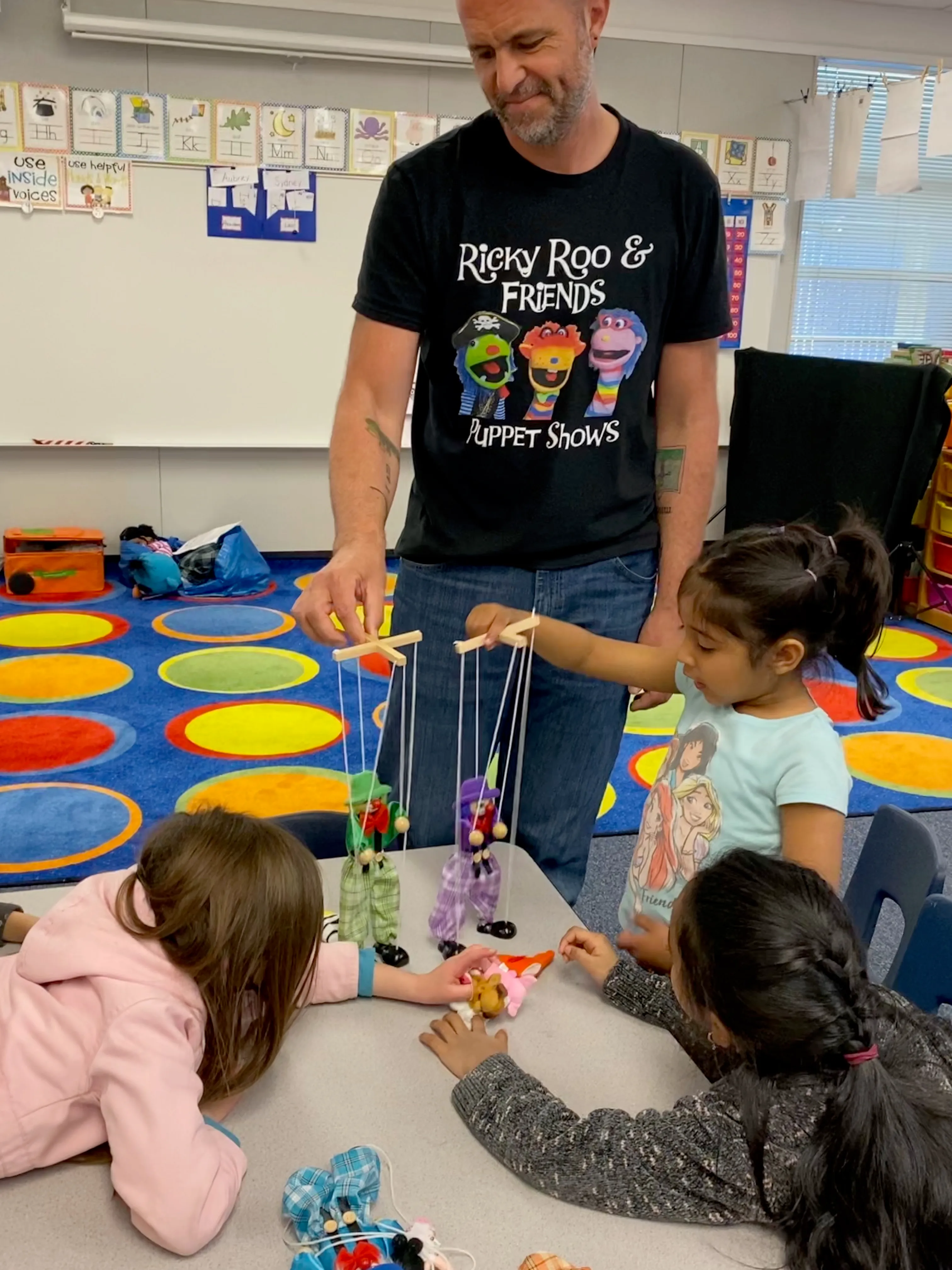Chris Rachowicz, founder of Ricky Roo & Friends Puppet Shows, teaching three children marionette puppeteering in a colorful classroom.