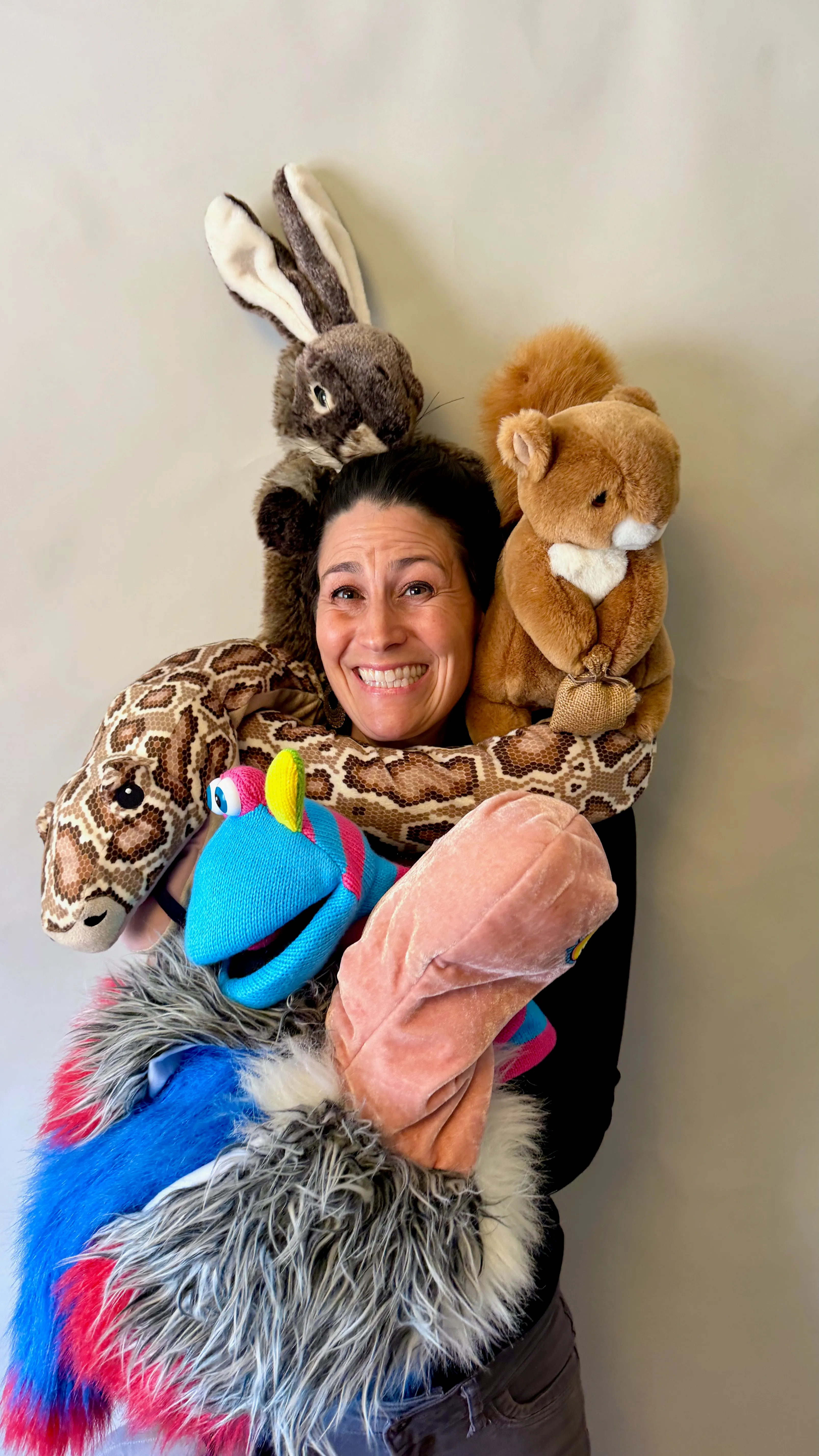 Smiling woman, puppeteer from RIcky Roo & Friends Puppet Shows, holding various colorful animal puppets including a snake, rabbit, squirrel, and blue bird.