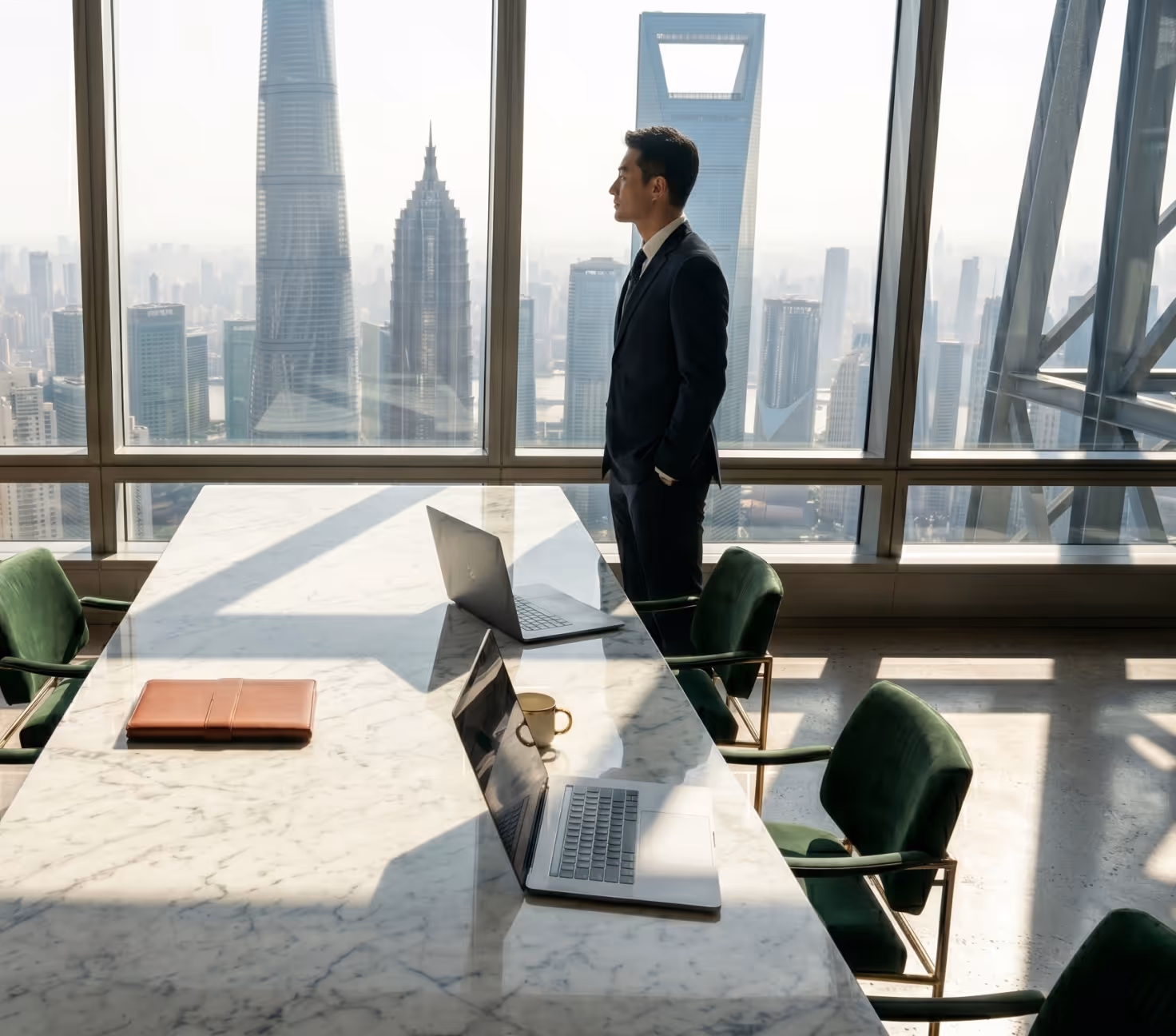 Businessman in suit standing at office window overlooking cityscape with skyscrapers and laptops on table.