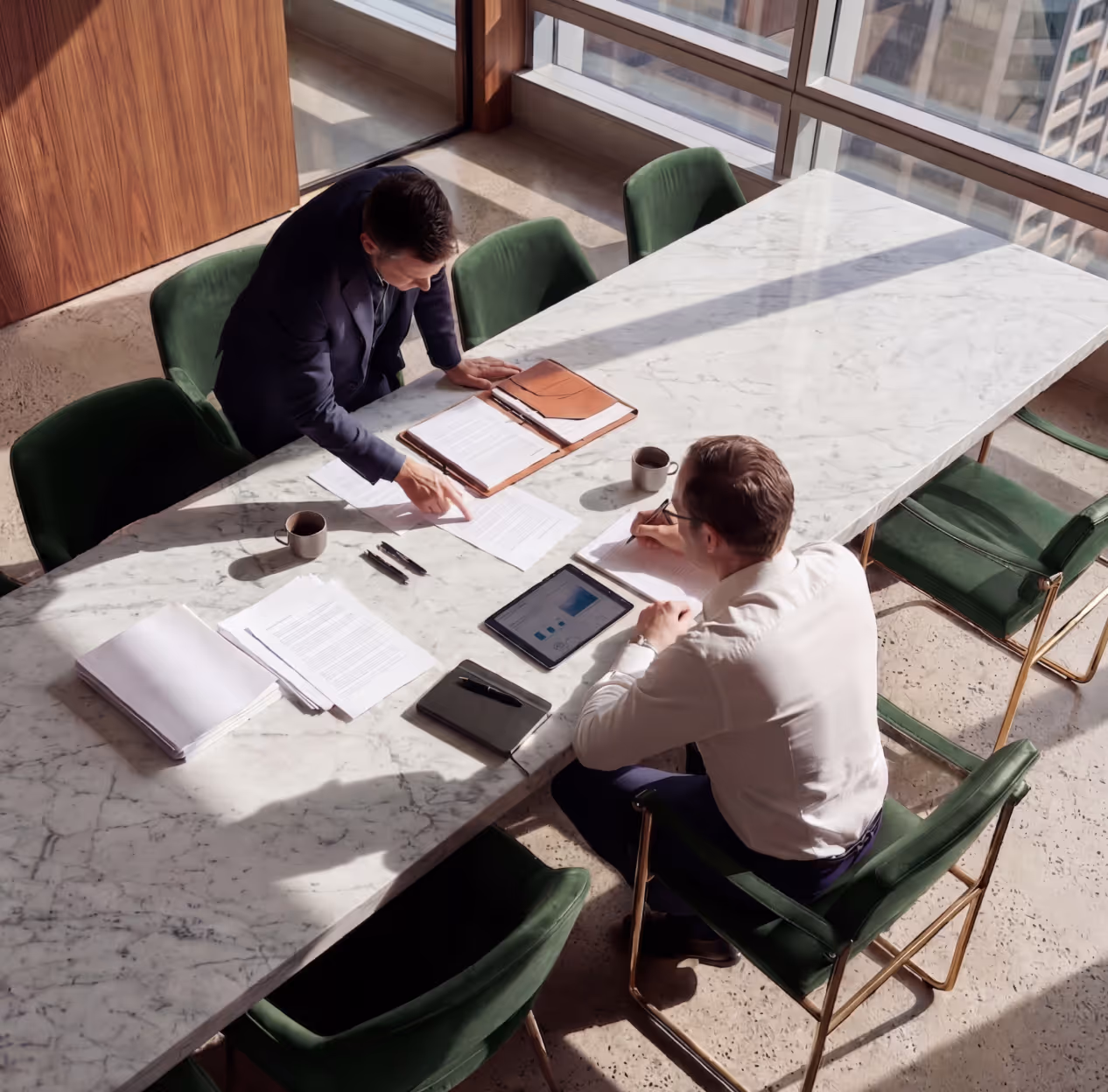 Two men in an office discussing documents at a marble table with an iPad and coffee cups.